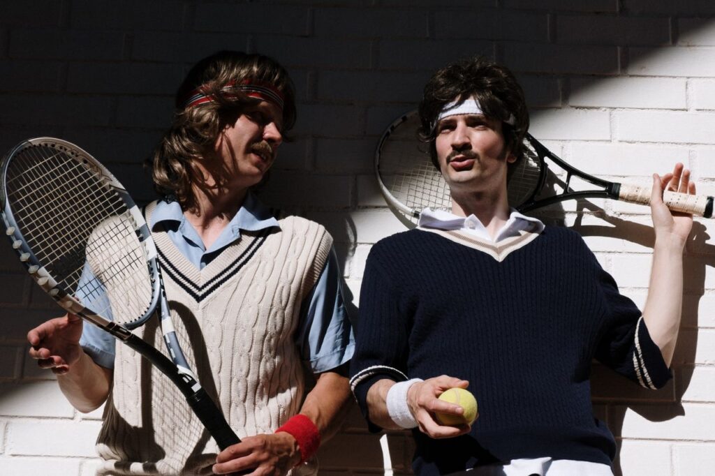 Two men in retro tennis attire with rackets and a tennis ball against a brick wall.